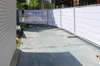 Ein Bauweg mit Metallplatten zwischen einem Gebäude und einem weissen Bauzaun, mit Bäumen im Hintergrund.
