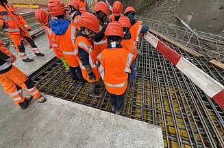 Eine Gruppe von Schulkindern in orangefarbenen Schutzwesten und Helmen steht auf Bewehrungsstahl auf einer Baustelle in Kloten und beobachtet die Bauarbeiten.