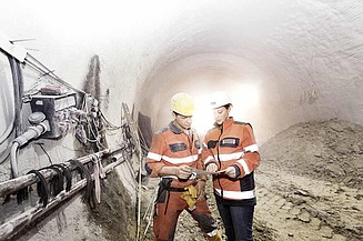 Zwei Bauarbeiter mit Helmen und Warnwesten stehen in einem hell beleuchteten Tunnel und prüfen Maschinen an der Tunnelwand.