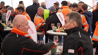 Menschen stehen dicht gedrängt in einem Festzelt an Stehtischen. Viele tragen orange STRABAG-Jacken oder dunkle Arbeitskleidung. Die Besucher unterhalten sich und halten Getränke in der Hand.