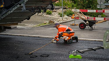 Ein Bauarbeiter verteilt heissen Asphalt mit einer Schaufel neben einer Strassenbaumaschine.