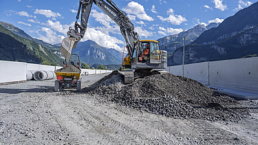 Ein Bagger verteilt Schotter auf einer Baustelle. Der Maschinenführer arbeitet auf einem grossen Steinhaufen, dahinter sind weisse Betonwände und die Berge zu sehen.