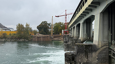 Baustelle am Flussufer: Sicht vom Wasser auf eine Staumauer, roten Kran und Spundwände der Baugrube.