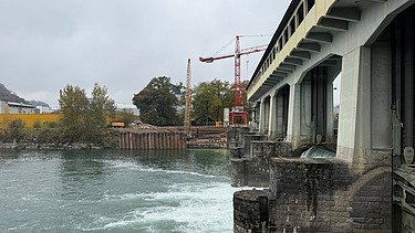 Baustelle am Flussufer: Sicht vom Wasser auf eine Staumauer, roten Kran und Spundwände der Baugrube.