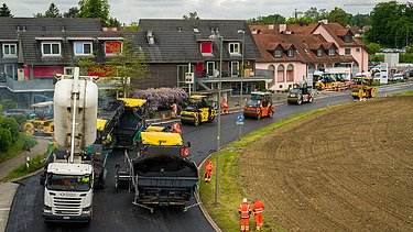 Baumaschinen und Arbeiter tragen frischen Asphalt auf eine Strasse in einem Wohngebiet auf.