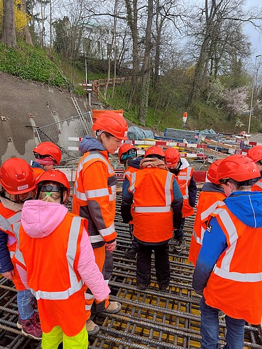 Schulkinder mit Schutzhelmen stehen auf Bewehrungsstahl und beobachten eine Baustelle in Kloten.