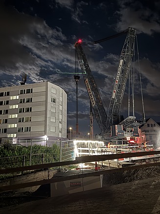 Eine nächtliche Baustellenszene zeigt zwei grosse Raupenkrane, die neben einem mehrstöckigen Gebäude arbeiten; helle Baustellenbeleuchtung reflektiert auf dem Boden, während im Vordergrund Maschinen und Material bereitstehen.