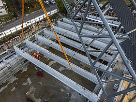 Mehrere grosse Turnhallenträger werden mit langen orangefarbenen Gurten von einem Kran in eine Halle gehoben.