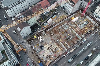 Luftaufnahme einer Baustelle mit einer grossen Baugrube inmitten von Stadtgebäuden. Bagger und Bauarbeiter führen Arbeiten im Tiefbau durch.