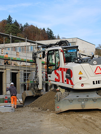 Bagger der STRABAG auf einer Baustelle vor einem Rohbau.