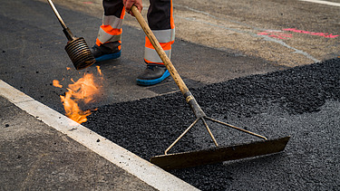 Ein Arbeiter glättet heissen Asphalt mit einem Werkzeug, daneben ist eine offene Flamme zu sehen.