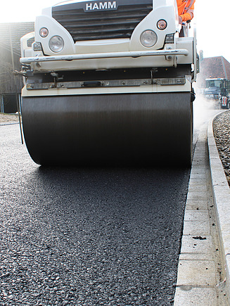  Eine weisse STRABAG Walze fährt an der Strassenkante und verdichtet den frischen dampfenden Asphalt.