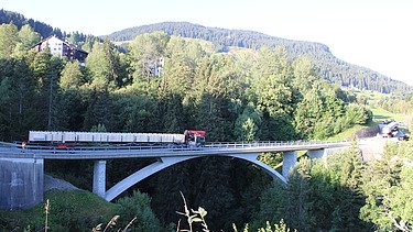 Ein langer Lastwagen fährt über eine hohe Brücke, die durch ein dicht bewaldetes Tal führt. Im Hintergrund sieht man steile, grüne Hänge und blauen Himmel.
