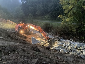 Arbeiten bei Dämmerung - STRABAG Ingenieurbau STRABAG Bagger arbeitet bei Abenddämmerung im Scheinwerferlicht am Aclettbach in Disentis. Im Hintergrund kann begrünte Bäume.