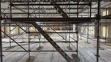 Innenaufnahme vom Landhaus mit einem umfangreichen Gerüstsystem und einer zentralen Treppe aus Metall. Der Boden ist mit Schutzfolie abgedeckt, auf der „MAXIMUM“ aufgedruckt ist. Das Gerüst reicht bis zur Decke und ermöglicht Arbeiten auf mehreren Ebenen im grossen, lichtdurchfluteten Raum mit hohen Fenstern.