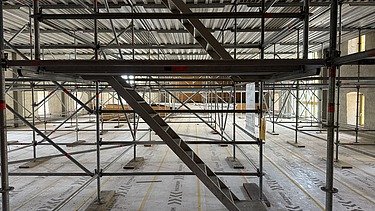Innenaufnahme vom Landhaus mit einem umfangreichen Gerüstsystem und einer zentralen Treppe aus Metall. Der Boden ist mit Schutzfolie abgedeckt, auf der „MAXIMUM“ aufgedruckt ist. Das Gerüst reicht bis zur Decke und ermöglicht Arbeiten auf mehreren Ebenen im grossen, lichtdurchfluteten Raum mit hohen Fenstern.