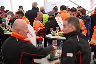 Menschen stehen dicht gedrängt in einem Festzelt an Stehtischen. Viele tragen orange STRABAG-Jacken oder dunkle Arbeitskleidung. Die Besucher unterhalten sich und halten Getränke in der Hand.