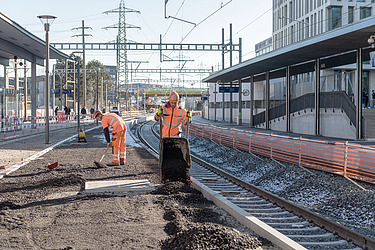 Zwei STRABAG - Mitarbeiter führen am Bahnhof Gleisbauarbeiten durch. Sie verteilen Schotter und bereiten den Untergrund für neue Gleise vor. Im Hintergrund sind Bahnsteige und Oberleitungen zu sehen.