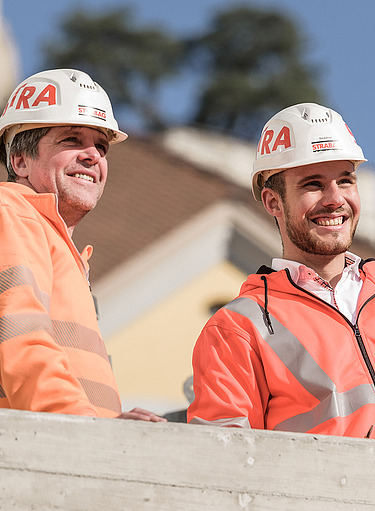 Zwei STRABAG-Mitarbeiter in oranger Arbeitskleidung und Helmen stehen auf einer Baustelle und schauen lächelnd zur Seite.