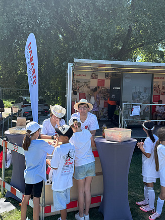 Kinder interagieren an einem Stand zwei KV-Lernenden vor dem mobilen Informationswagen von STRABAG.
