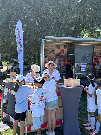 Kinder interagieren an einem Stand zwei KV-Lernenden vor dem mobilen Informationswagen von STRABAG.