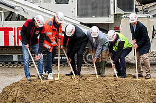 Mehrere Personen in Schutzkleidung und Helmen heben beim Spatenstich gemeinsam Erde aus. Im Hintergrund ist schweres Baustellengerät zu sehen.