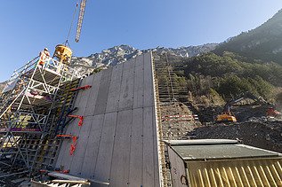 STRABAG-Baustelle im Gebirge: Betonarbeiten an einer hohen Stützmauer mit Kran und Bauführer:in auf dem Gerüst vor Bergkulisse.